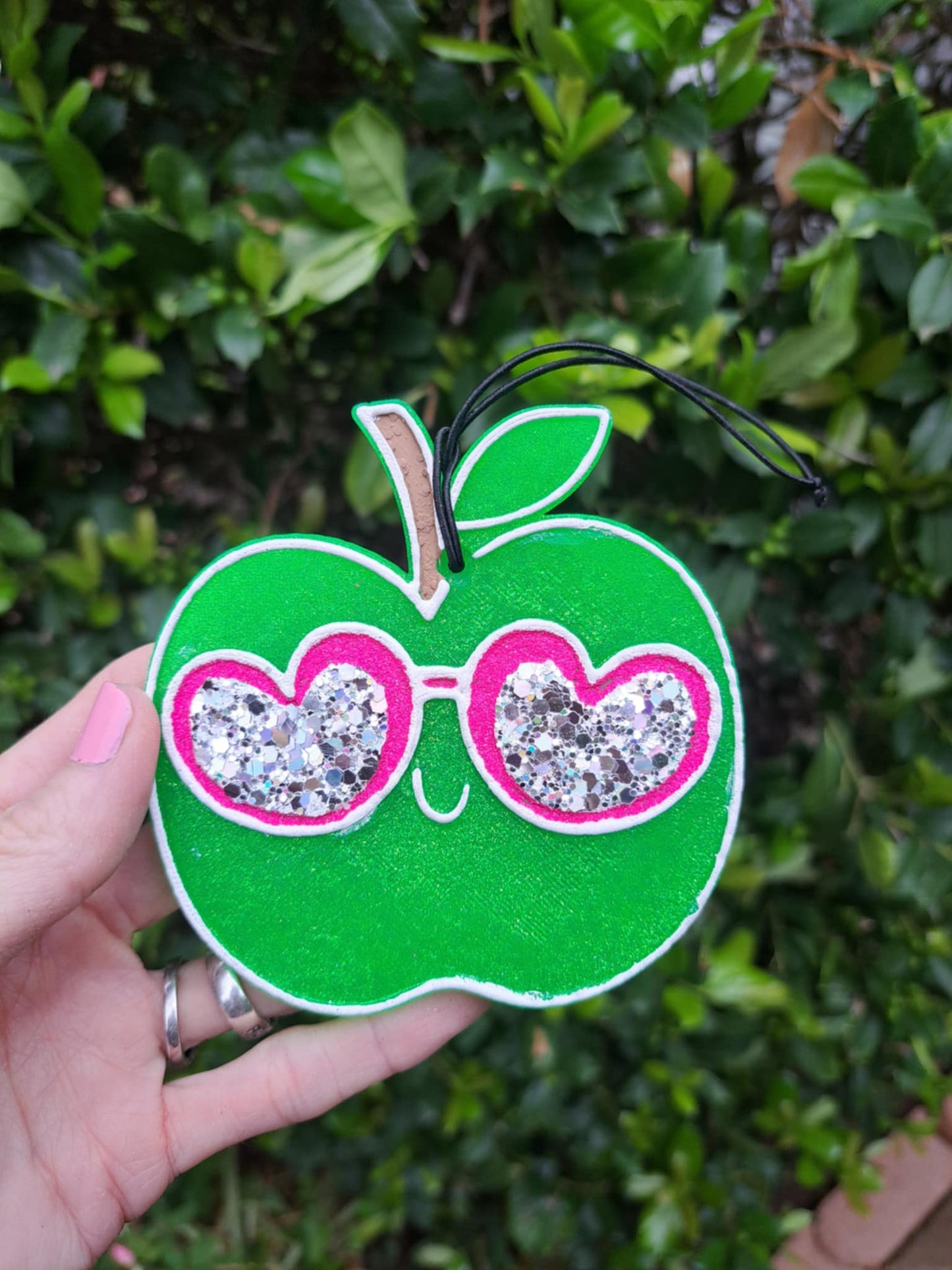 Handmade green apple-shaped air freshener with a design of sunglasses on it, held in a hand with a foliage background.