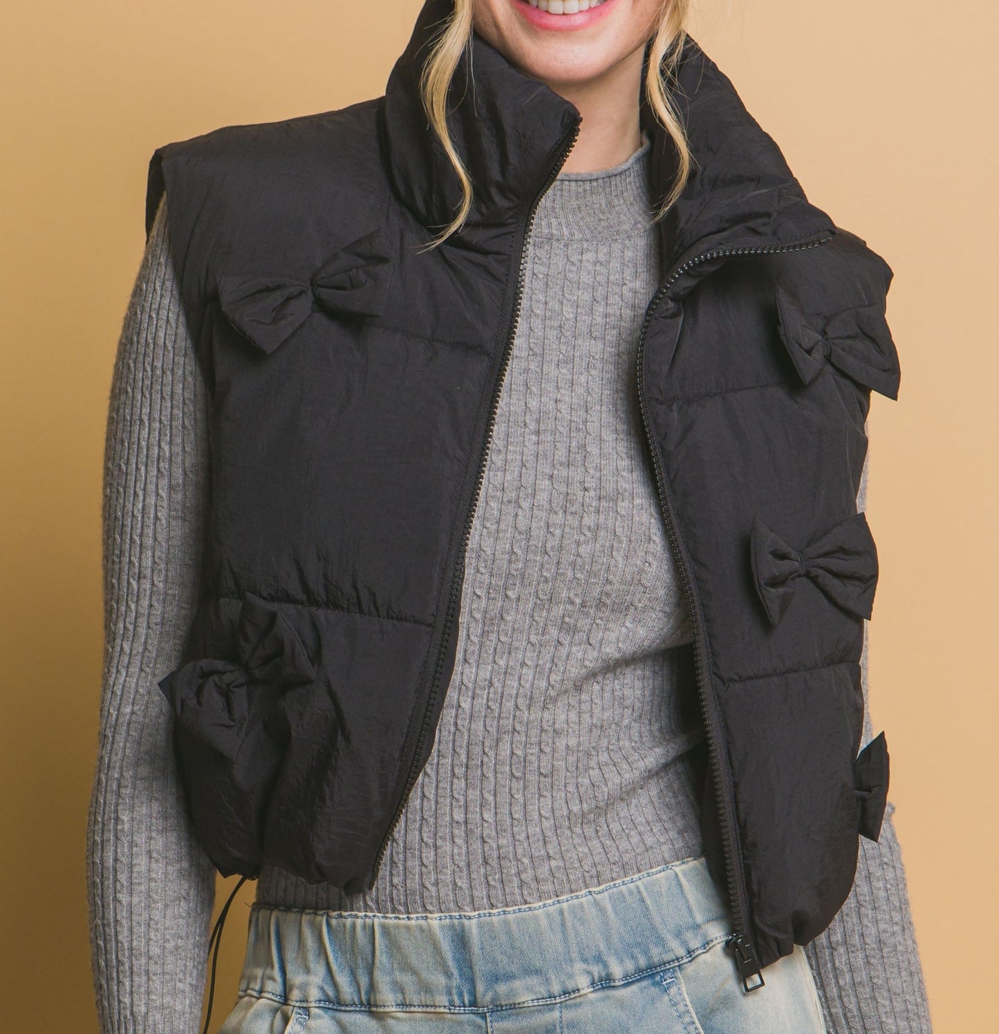 A woman wearing a black zip front puffer vest with bow details on the front, over a grey sweater, with jeans and a grey top.
