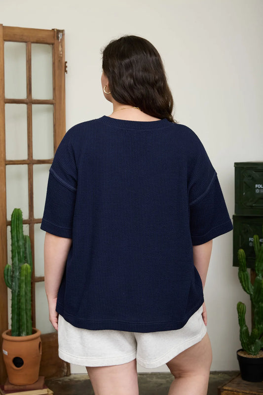 Person wearing a navy blue shirt and white shorts standing in a room with plants.