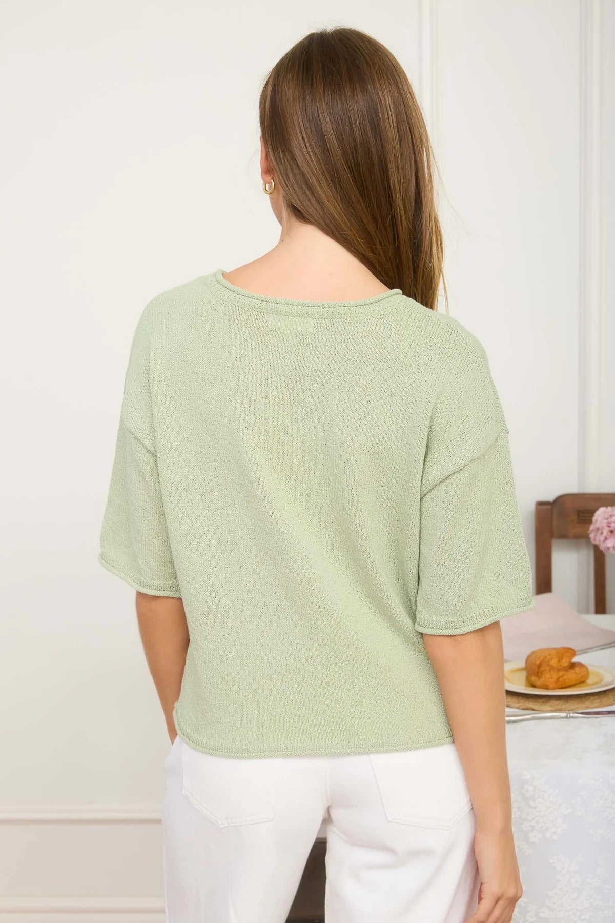 Woman wearing a light green top and white pants, standing in a room with a table and chair.