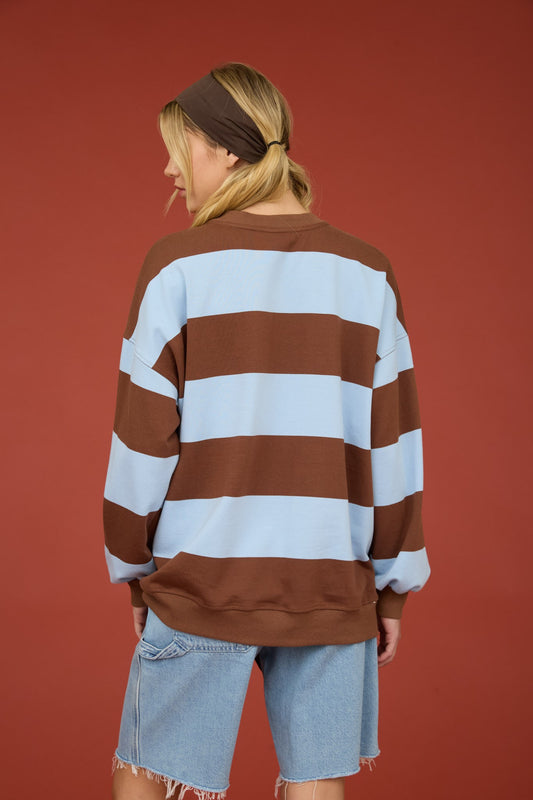 A woman shown from the back wearing an oversized long-sleeve sweatshirt with horizontal brown and light blue stripes, paired with light-wash denim shorts, against a solid terracotta red background.