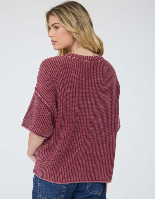 A woman shown from the back wearing a ribbed knit short-sleeve sweater in an acid washed burgundy color paired with blue jeans, against a plain light gray background.