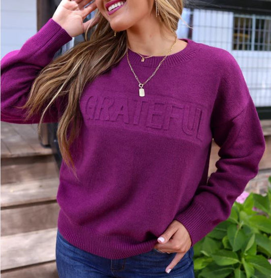 A woman wearing a plum purple embossed sweater with the word 'grateful' on the front, paired with blue jeans and a necklace.