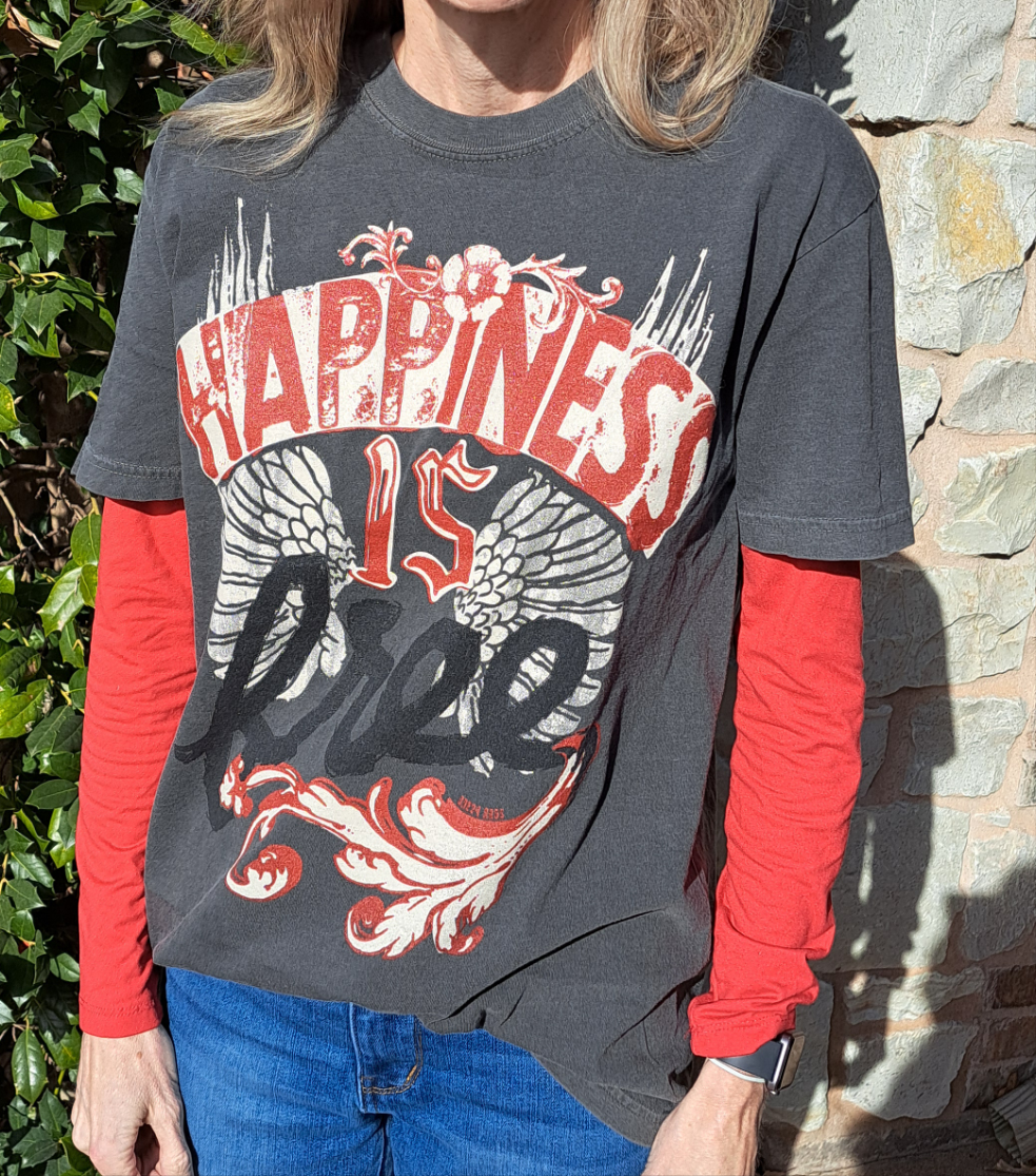 A person standing outdoors wearing a charcoal gray t-shirt with the text 'Happiness is FREE' printed on the front, paired with red long-sleeve shirts and blue jeans.