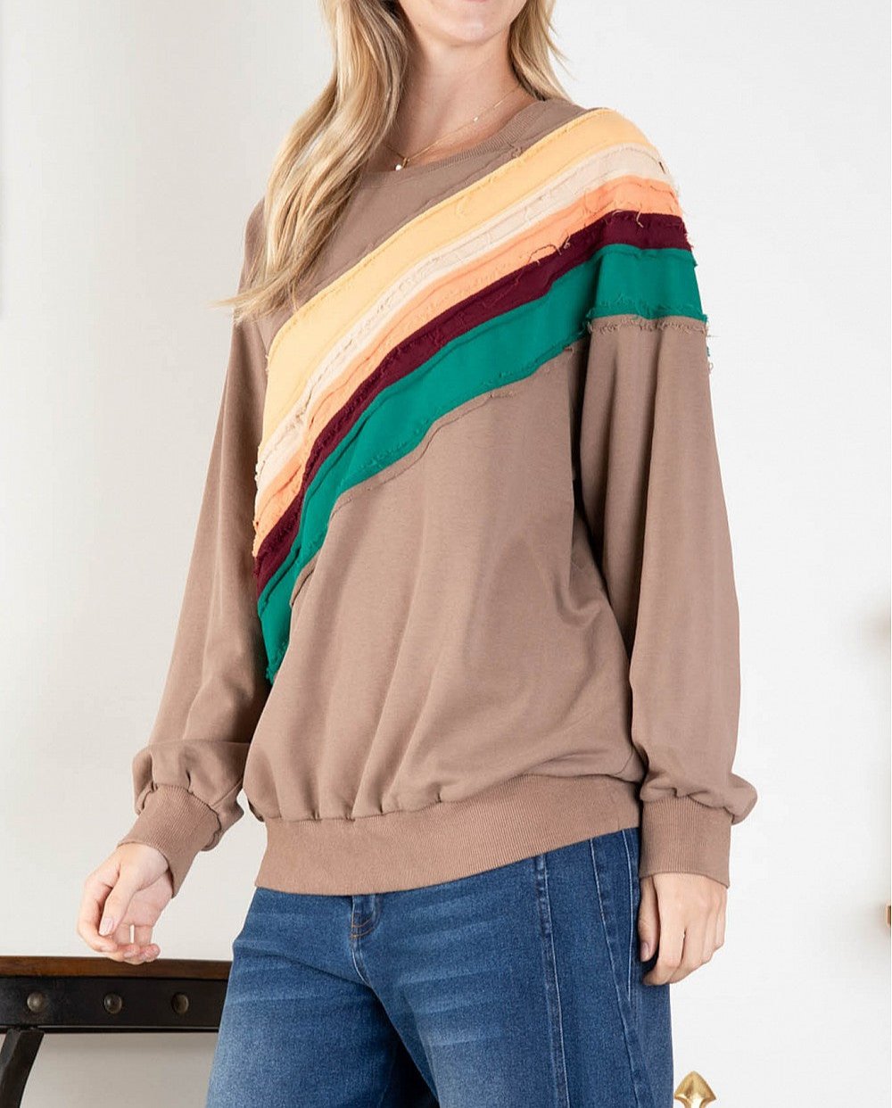 A woman wearing a mocha brown sweatshirt with a fall rainbow stripe pattern, long sleeves, and a ribbed cuff around the neck, wrists, and bottom of the garment. She is also wearing blue jeans.