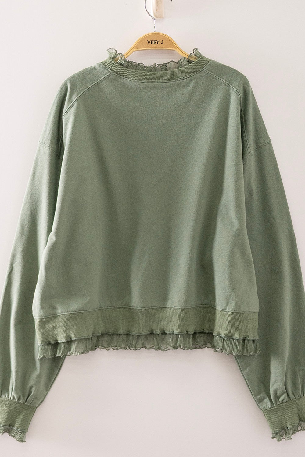 An olive green long-sleeve sweatshirt with ruffled trim details, hanging on a wooden hanger against a plain white wall.