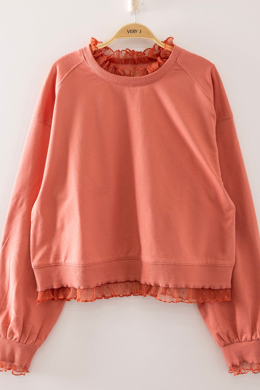 A coral pink long-sleeve sweatshirt with a ruffled mock neck and ruffled hem, hanging on a wooden hanger against a plain white wall.