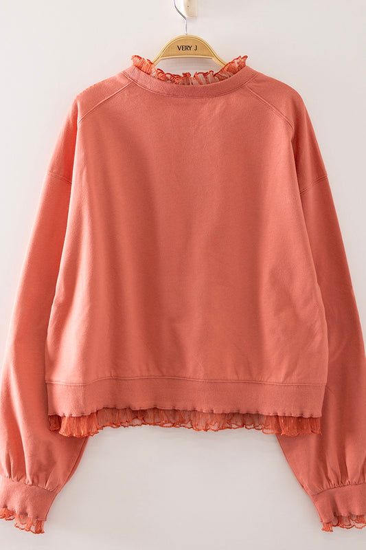 A coral pink long-sleeve sweatshirt with a ruffled mock neck and hem, hanging on a wooden hanger against a plain white wall.