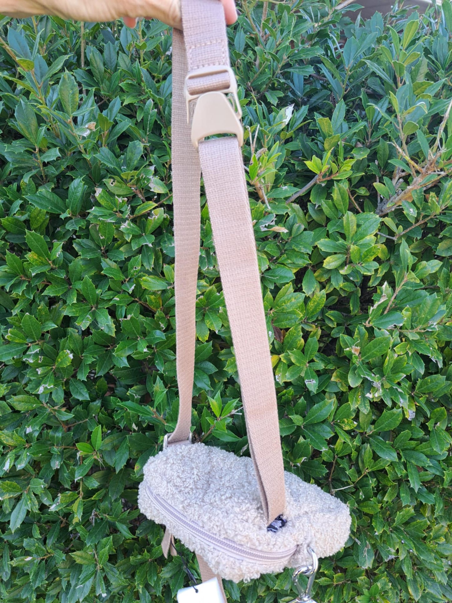 Small white bag with a beige belt strap against a green bush background