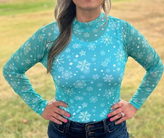 A woman wearing a turquoise mesh top with white snowflake print, paired with blue jeans. Green background