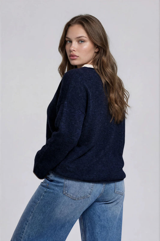 A woman wearing a dark navy blue knit sweater and light blue jeans, photographed from the back against a plain light gray studio background.