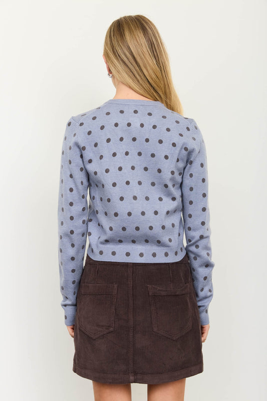 A woman shown from the back wearing a light blue long-sleeve knit top with a dark polka dot pattern and a dark brown mini skirt, against a plain off-white background.