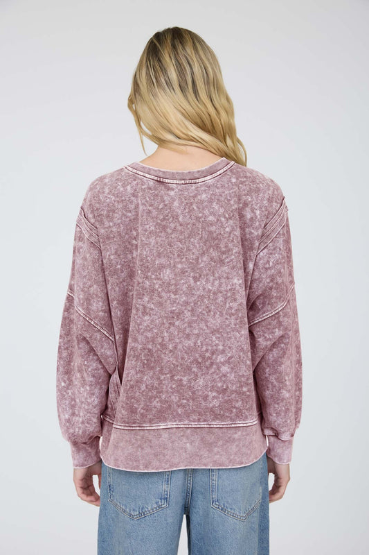 A woman viewed from the back wearing a dusty rose acid-wash sweatshirt and light blue jeans against a light gray studio background.