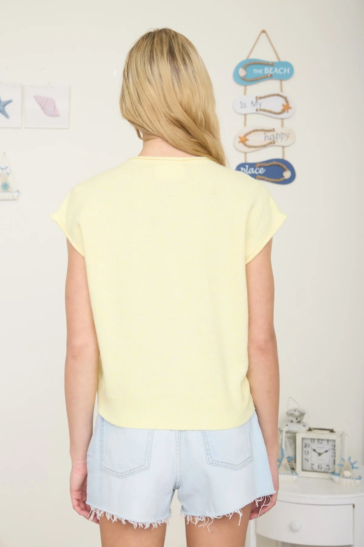 Back view of a person wearing a yellow top and denim shorts in a room with decorative items on the wall.