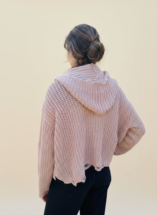 back view of a person wearing a blush pink sweater hoodie and black pants standing in front of a cream background