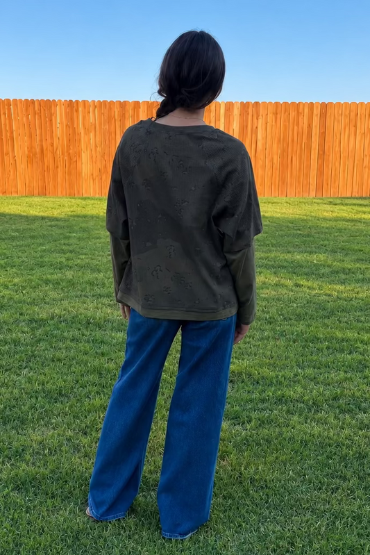 Back view of a lady wearing an olive layered camo top with jeans standing in grass with a fence behind her