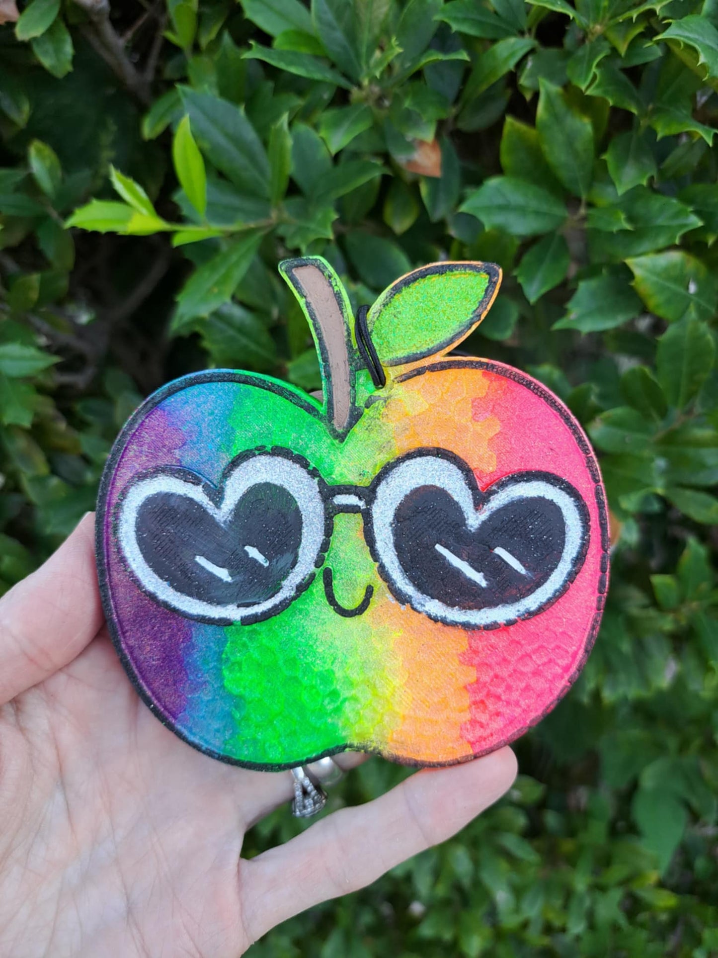 Colorful apple freshie with heart sunglasses held in front of green foliage.
