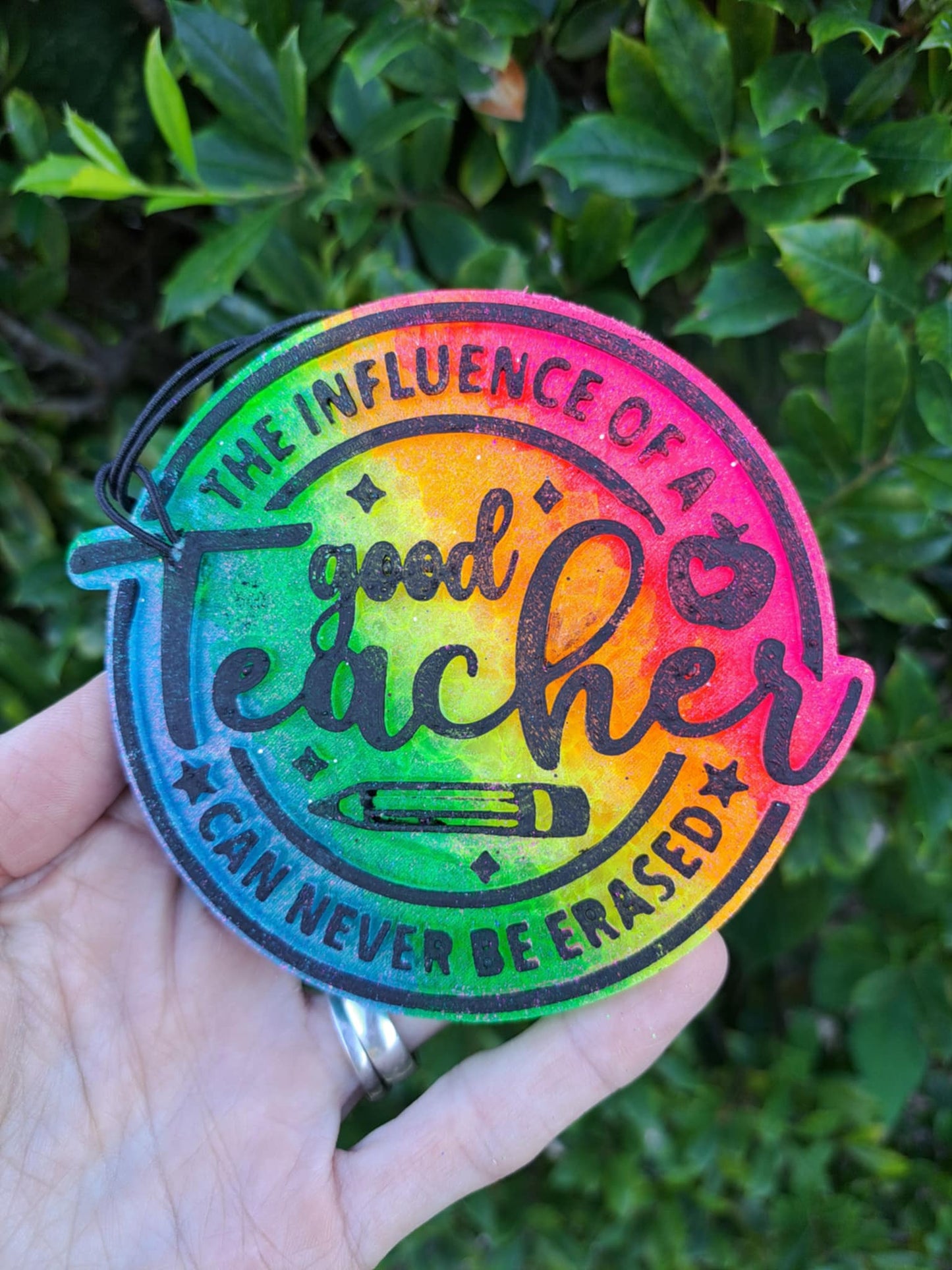 Colorful freshie with black text 'good teacher' held by a hand against a green leafy background