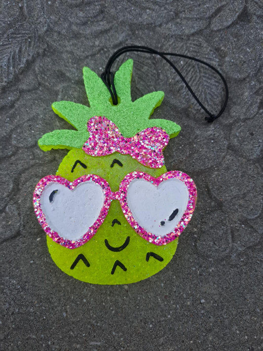 Pineapple-shaped freshie with pink glittery bow and heart-shaped glasses on a textured gray background