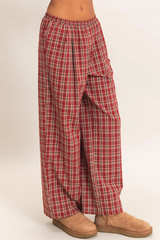 Red and white plaid lounge pants with a wide elastic waistband and side cargo pocket, shown on a model wearing tan suede slippers against a light beige background.