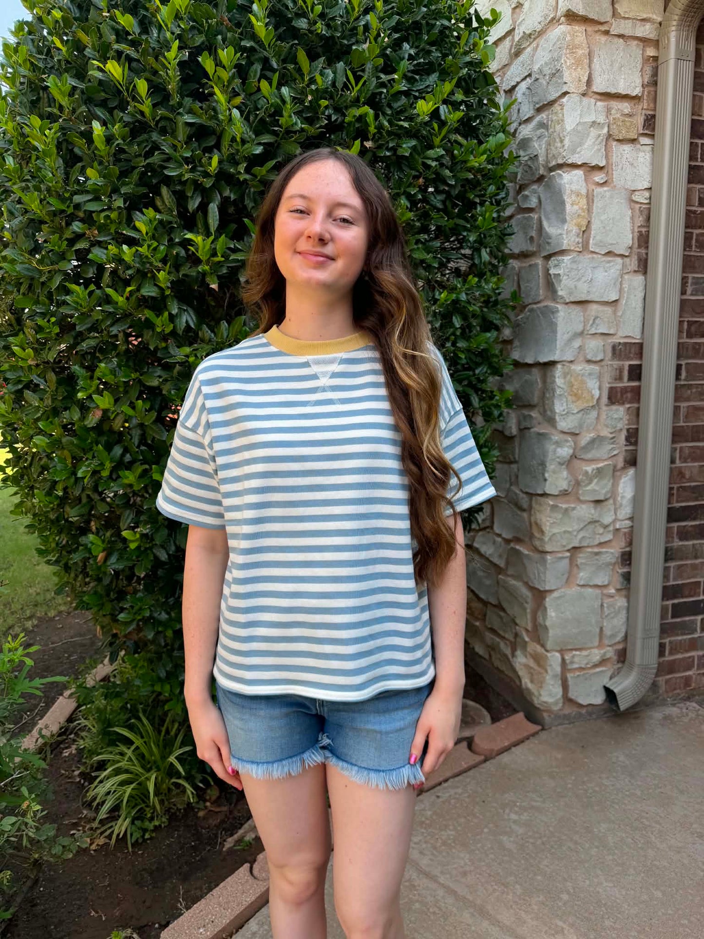 A young woman stands outdoors in front of a stone wall and green hedge, wearing a light blue and white striped short-sleeve t-shirt with a yellow collar trim and denim shorts.