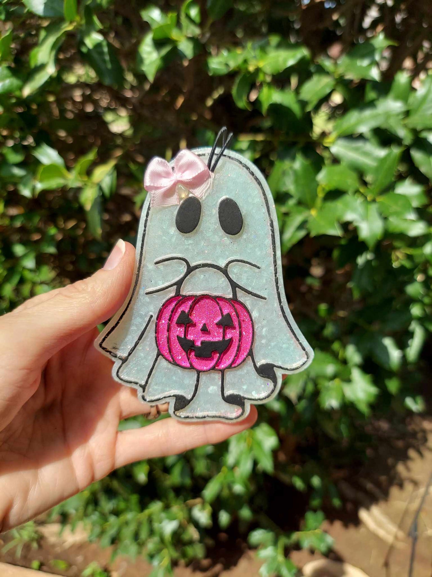 A hand holding a small air freshener shaped like a ghost with a pink pumpkin, against a backdrop of greenery.