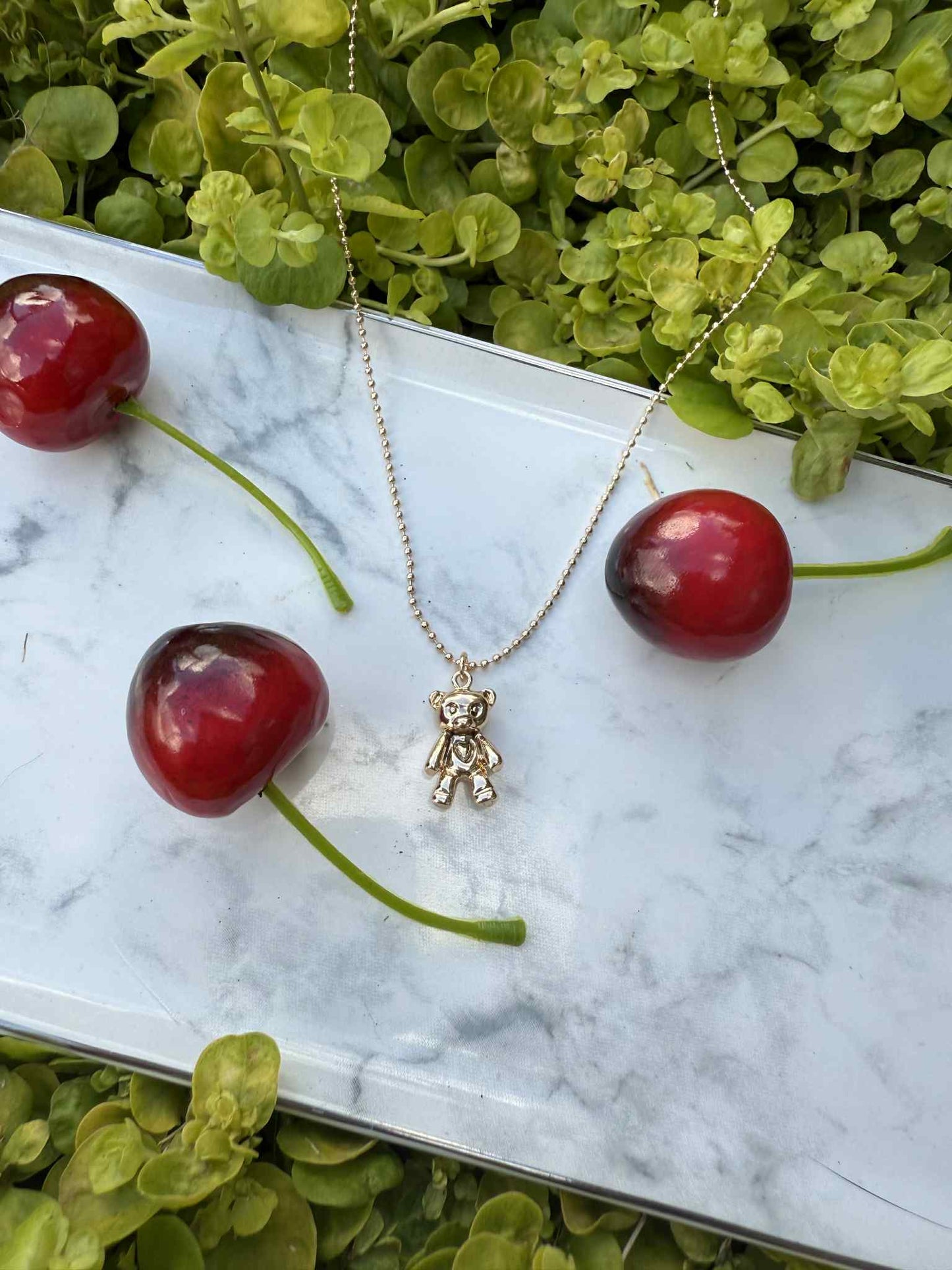 Gold teddy bear necklace on a marble surface with cherries and green leaves.