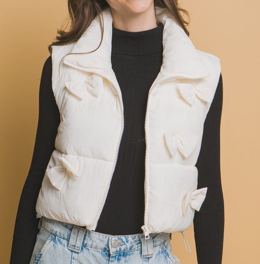 A woman wearing a black top and blue jeans, with a zip front ivory puffer vest with bow details on the front and at the waist, and adjustable drawstrings.