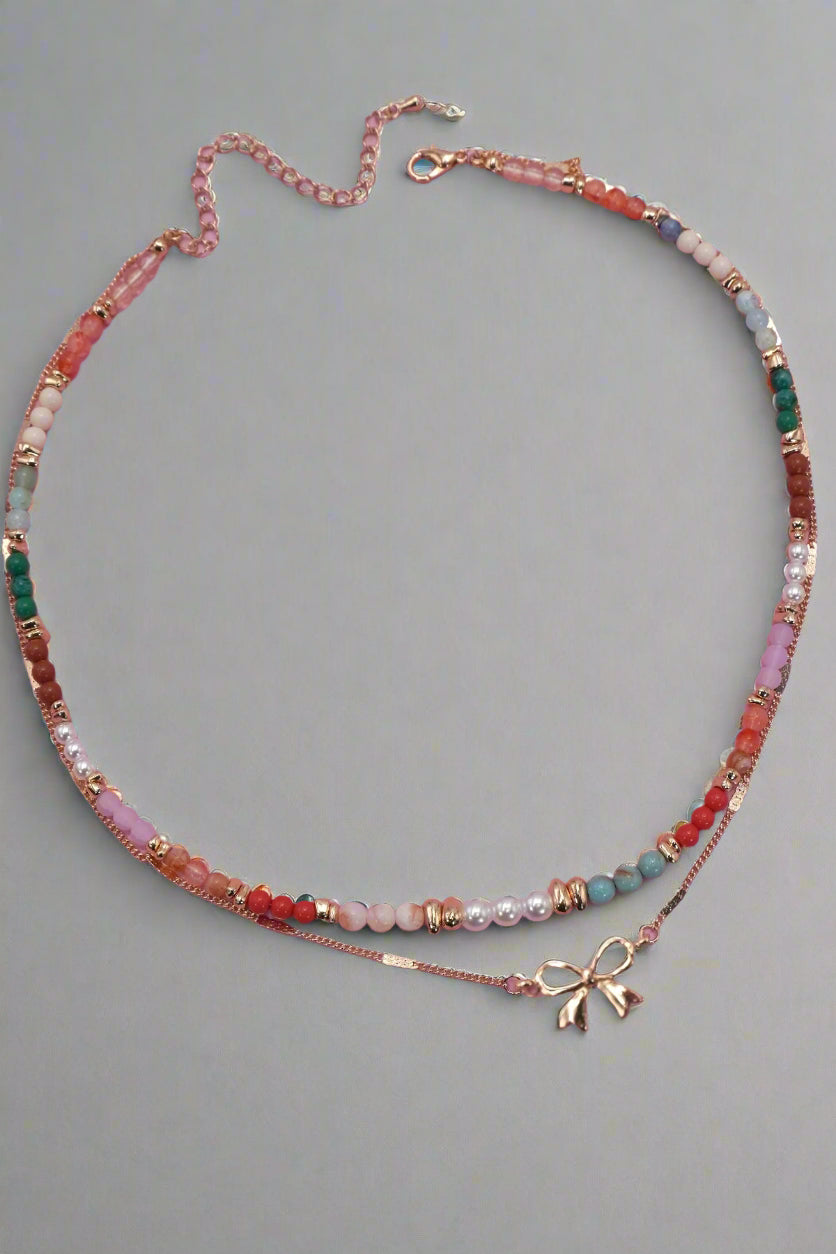 A multi-beaded, gold-plated double layer necklace with a bow charm on a pink background.