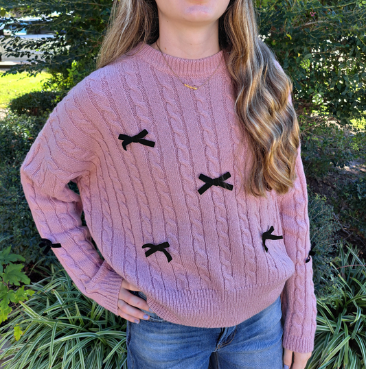 A person wearing a pink cable knit sweater with black velvet bows on the front.