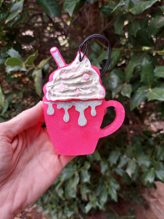 A hand holding a pink mug-shaped air freshener with whipped cream and candy sprinkles design, against a backdrop of green foliage.