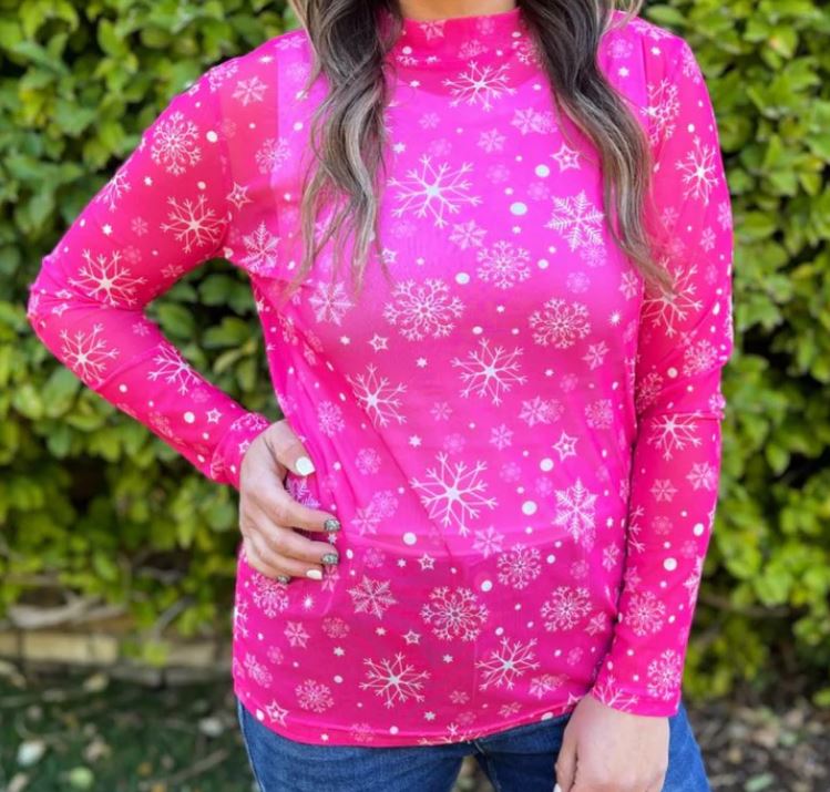 A woman wearing a hot pink mesh top with white snowflake print, standing in front of greenery