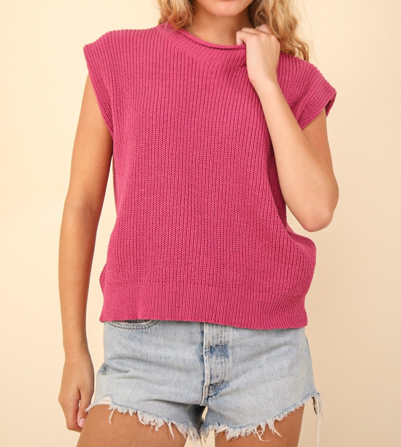 A female model wearing a pink oversized knit sweater vest top with a round neckline and ribbed hem, paired with denim shorts.