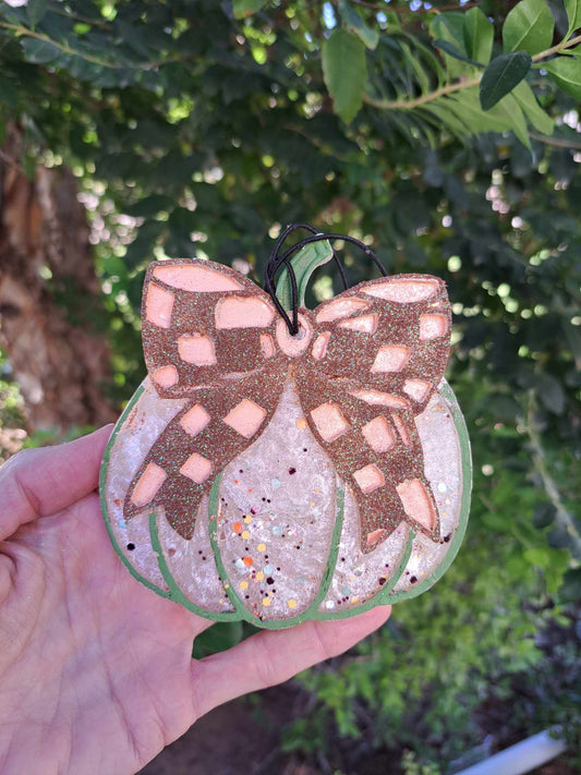 A hand holding a pumpkin-shaped air freshener with a camel check bow design, against a backdrop of greenery.