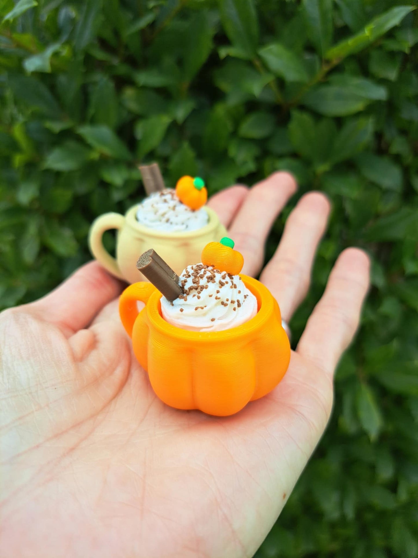 Two small pumpkin-shaped cups with whipped cream and a cinnamon stick held in a hand against a green leafy background.