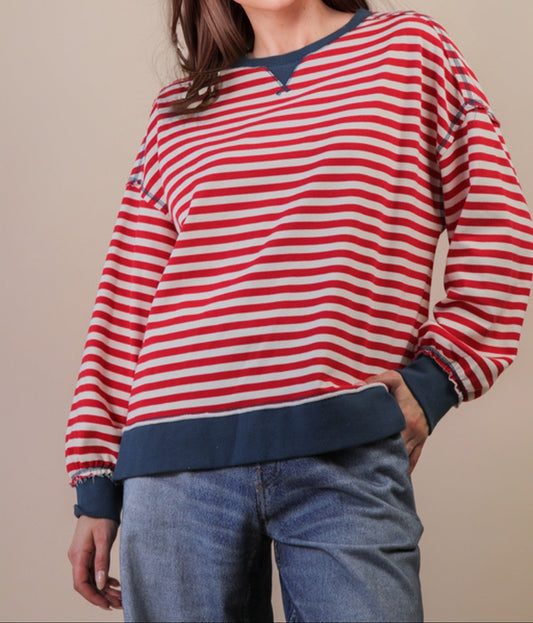 red and cream knit long sleeve top with blue trim on person wearing jeans. Beige background
