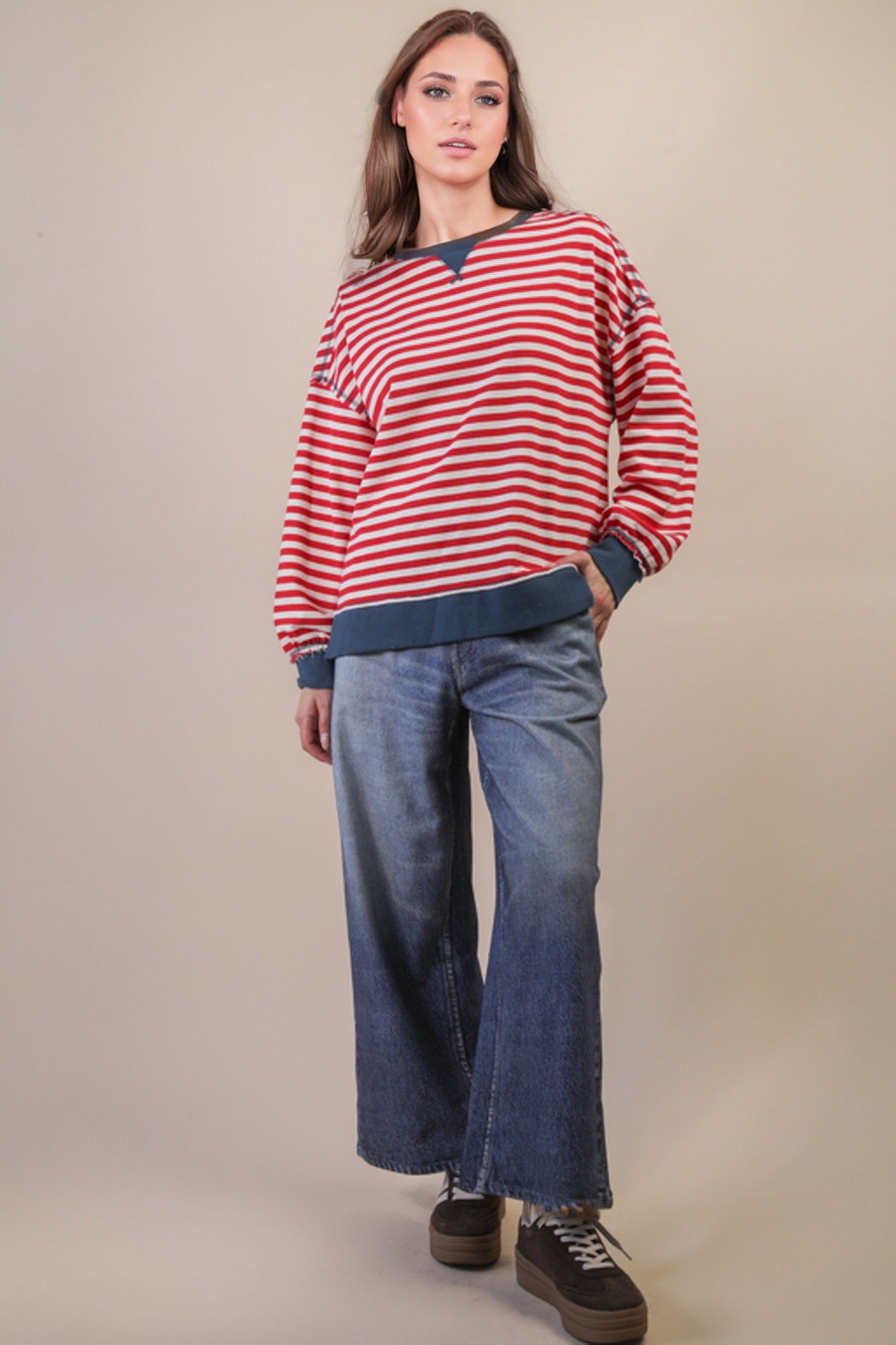 red and cream striped long sleeve top with blue trim on a person wearing jeans and brown sneakers. Beige background