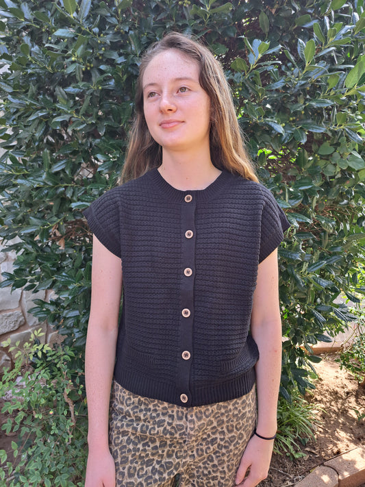 Person wearing a black knitted vest over a leopard print dress standing in front of green foliage.