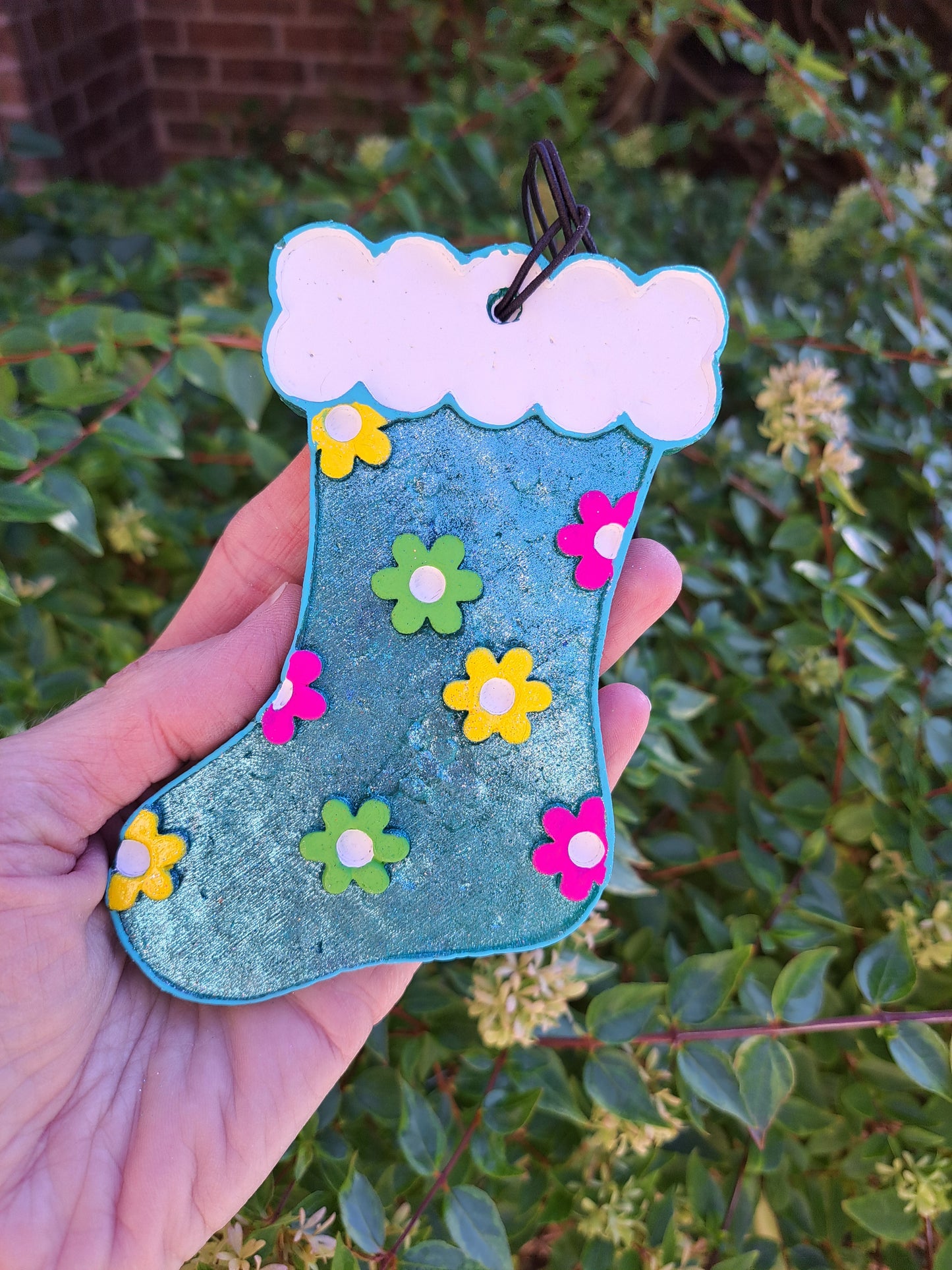 A hand holding a blue metallic air freshener shaped like a stocking with colorful flowers. Greenery background.