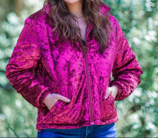 A woman smiling, wearing a wine colored lush velvet puffer jacket with a front zip closure and side pockets, standing in a natural outdoor setting.