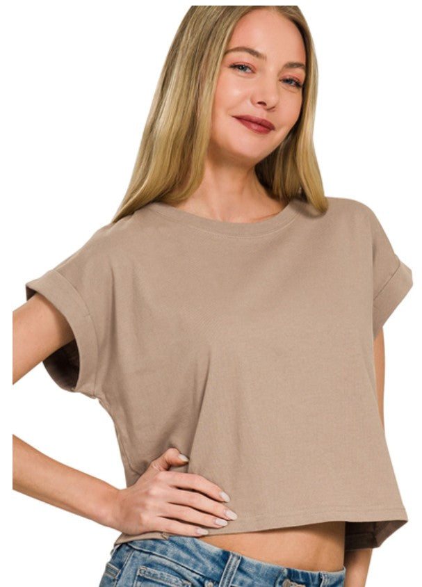 A woman wearing a cropped, short-sleeve t-shirt in a light taupe-beige color, paired with blue jeans, shown against a plain white background.