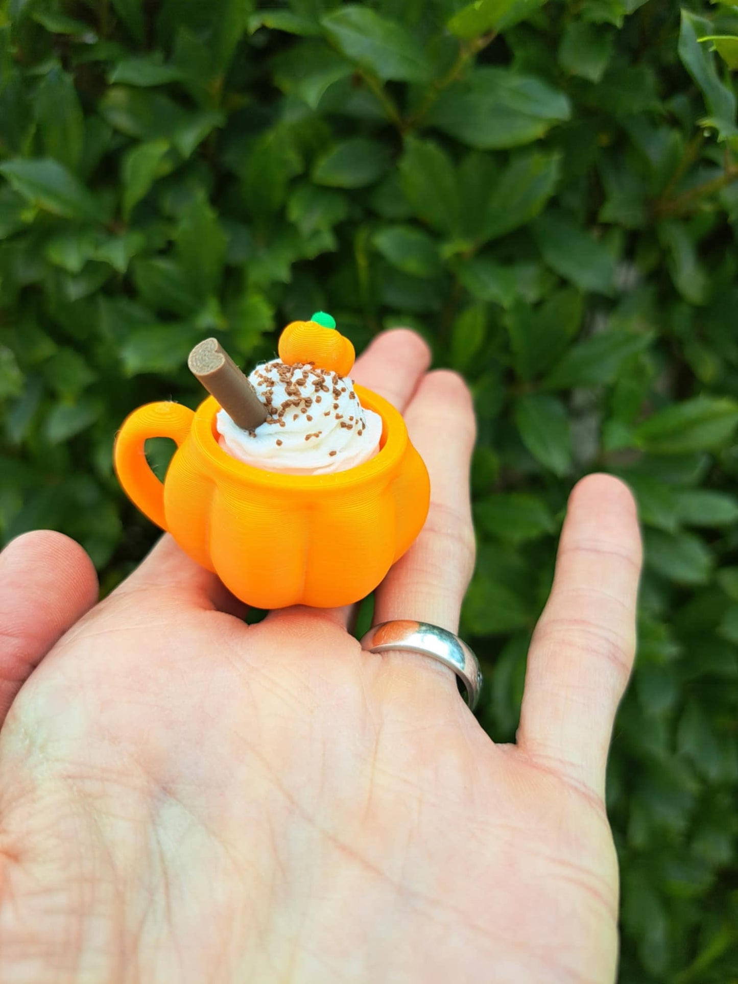 Pumpkin-shaped cup with a straw held in a hand against a green leafy background