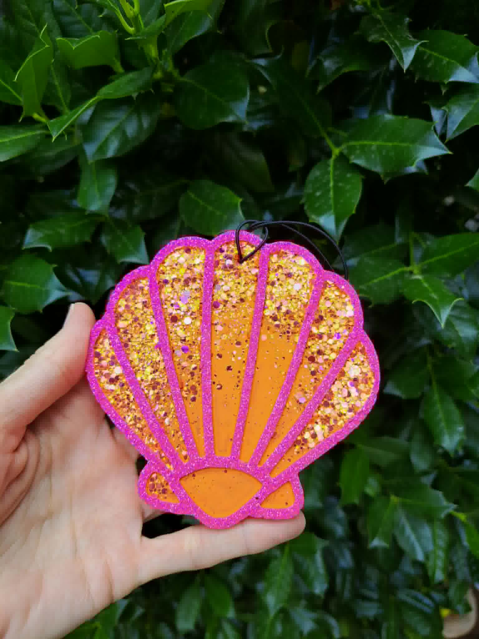 Hand holding a glittery shell-shaped freshie against a green leafy background