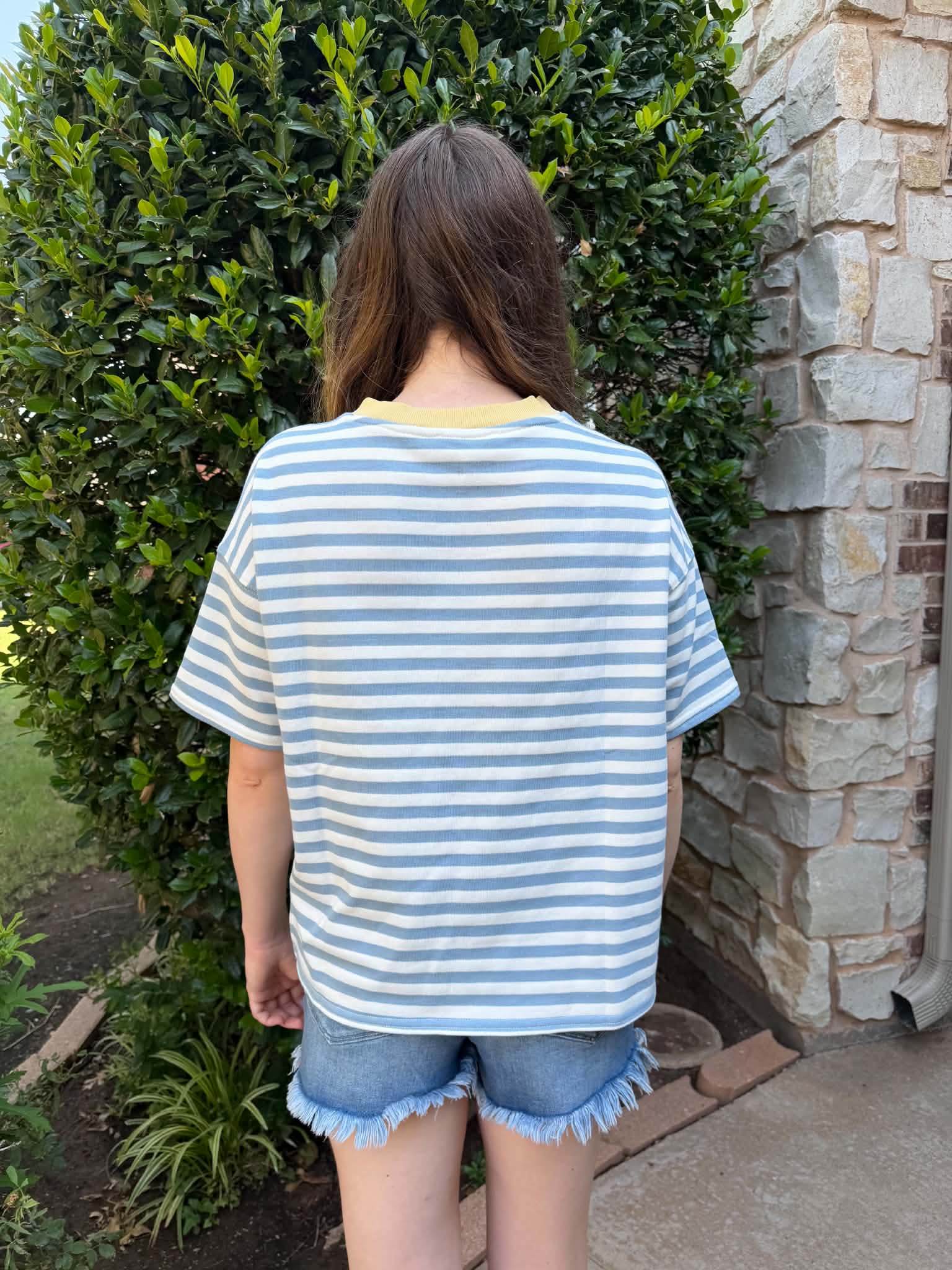 A person viewed from behind wearing a light blue and white striped t-shirt with a yellow inner collar, paired with denim shorts, standing outdoors in front of a stone wall and green hedge.
