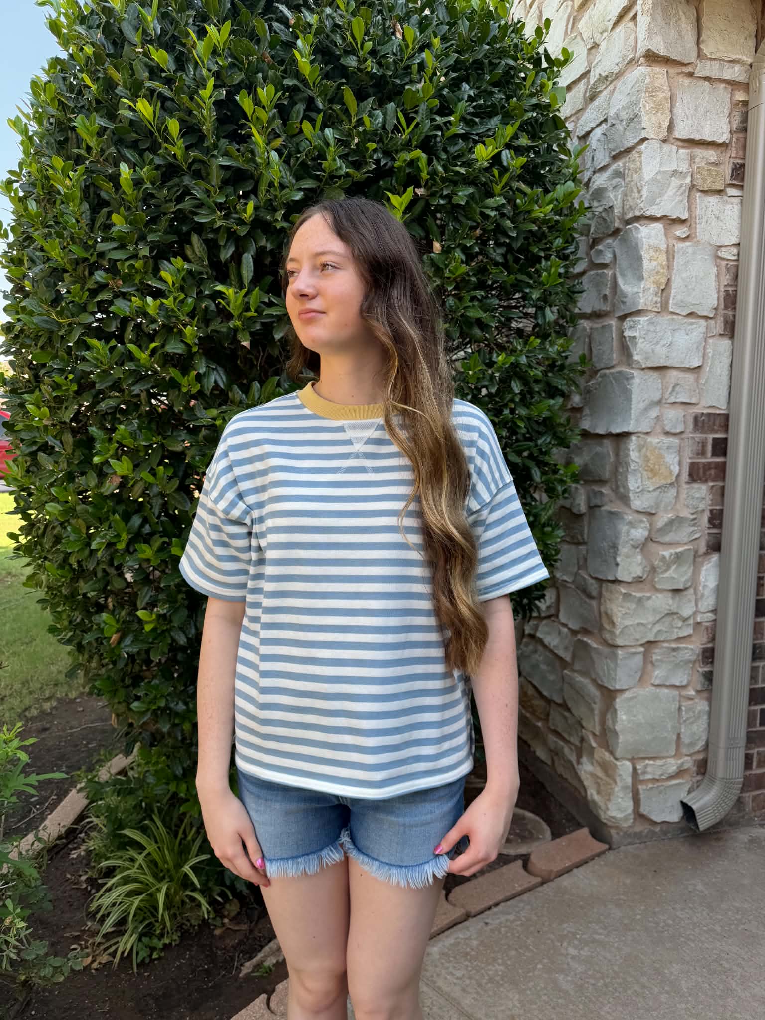 A young woman stands outdoors in front of a stone wall and green hedge, wearing a light blue and white striped short-sleeve t-shirt with a yellow crew neck, paired with denim shorts.