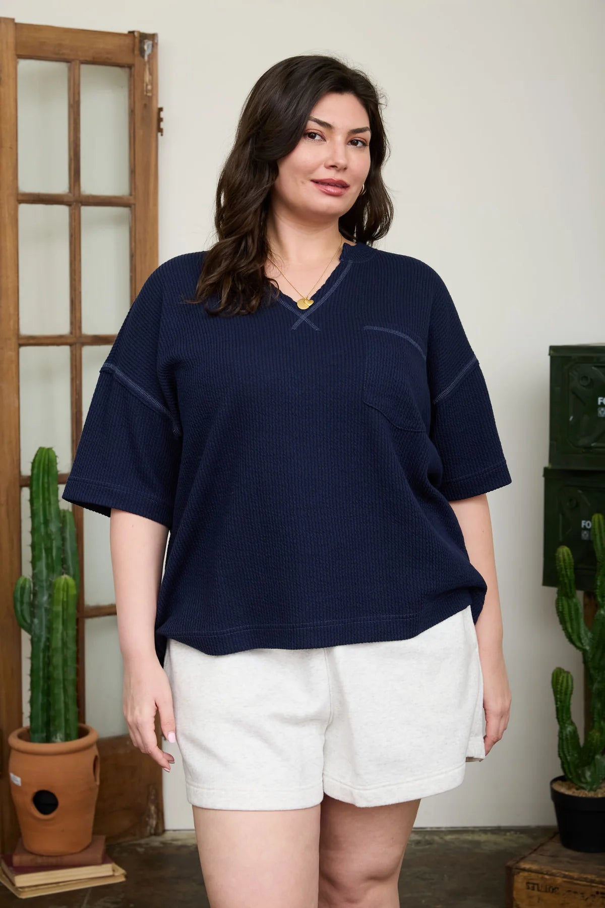 Woman wearing a navy blue top and white shorts standing indoors with plants in the background