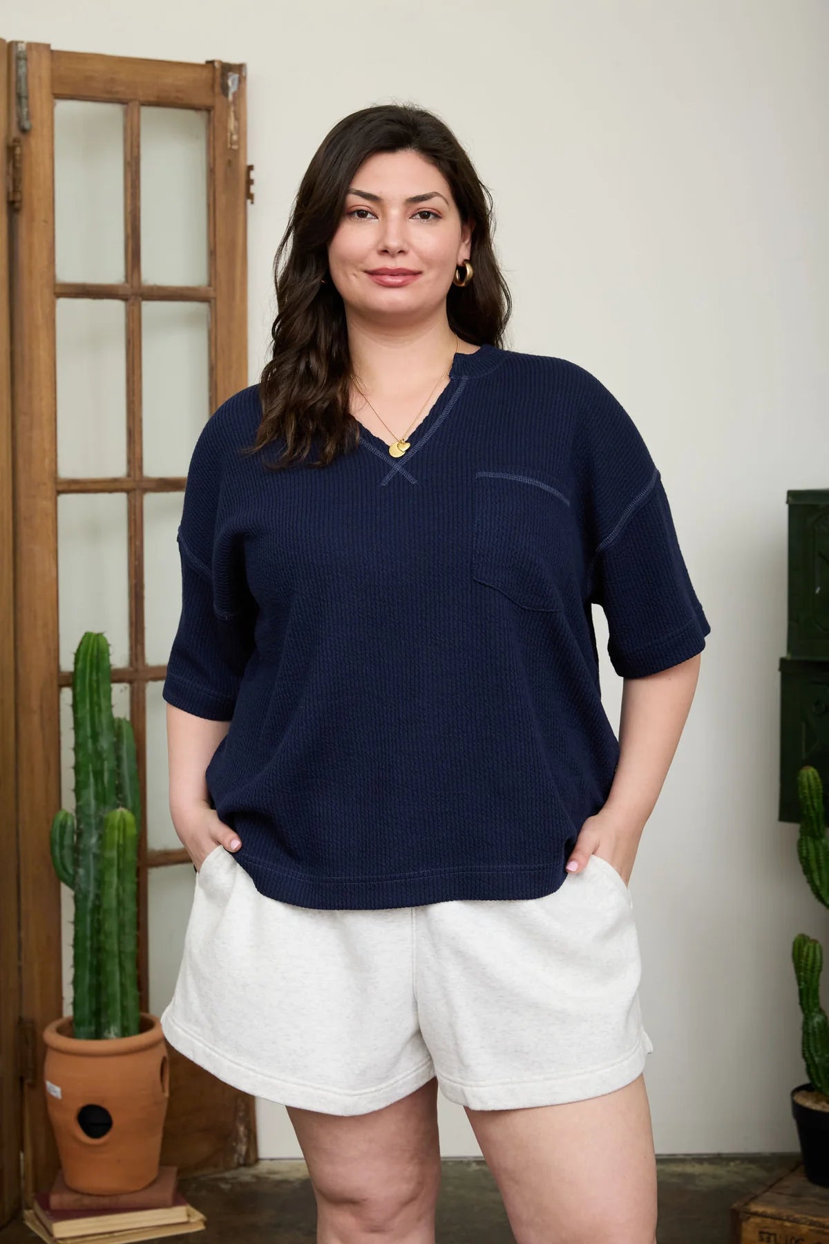 Woman wearing a navy blue sweater and white shorts standing indoors with plants in the background