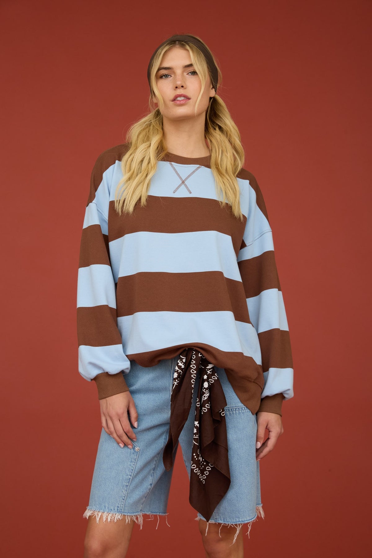 A woman modeling a brown and light blue horizontally striped long-sleeve sweatshirt with lace-up detail at the chest, paired with light-wash denim shorts and a dark brown patterned bandana tied at the waist, set against a solid terracotta background.