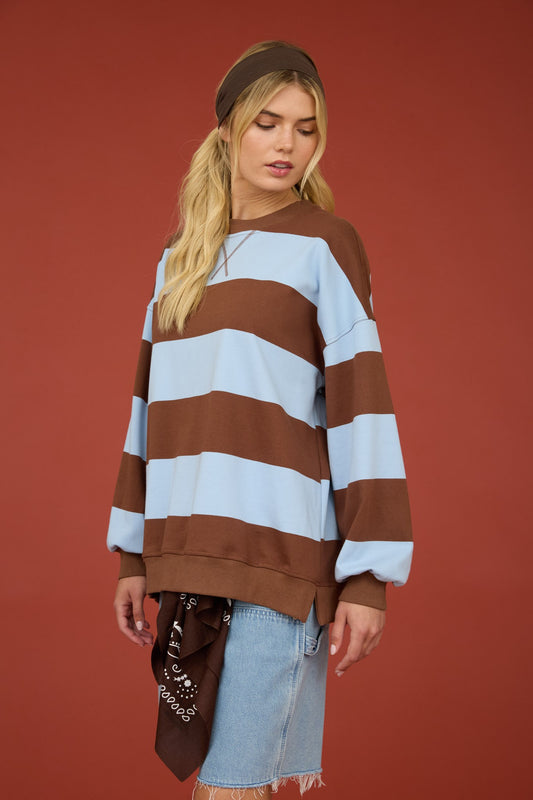 A woman wearing a brown and light blue horizontally striped sweatshirt, a brown beanie, and a denim skirt, standing against a solid terracotta red background.