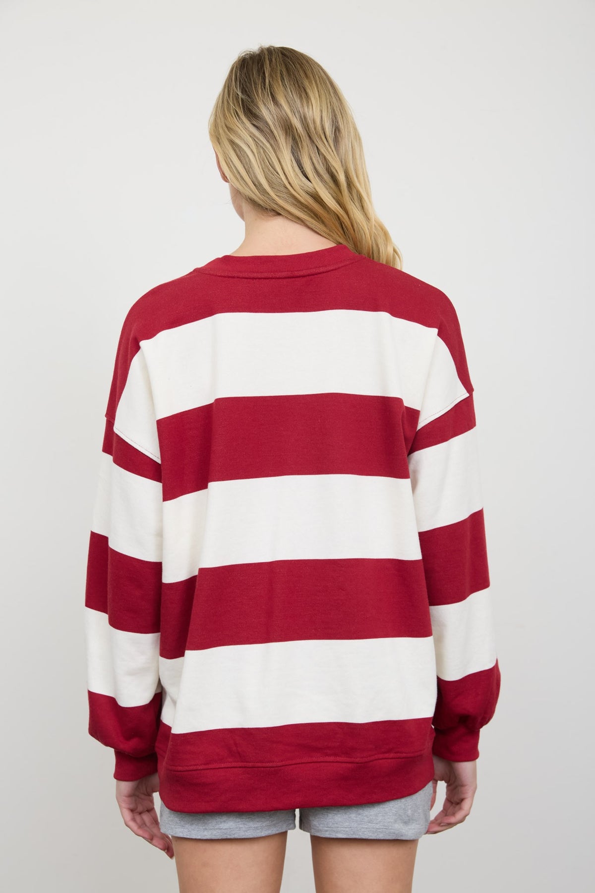 A woman viewed from behind wearing a red and white horizontally striped long-sleeve sweatshirt and gray shorts against a light gray studio background.